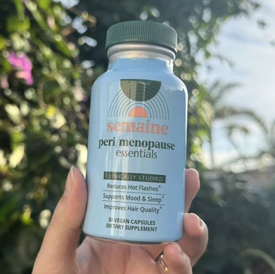 A woman's hand holds a light blue bottle of peri/menopause essentials by Semaine Health. There is a blue sky and a tree blurred in the background