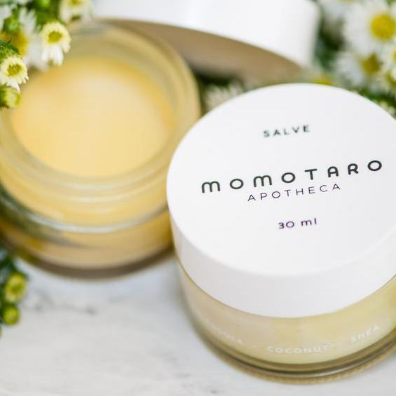 An open container of vaginal salve with the cap resting on it sits next to an unopened jar. They are surrounded by small white and yellow flowers