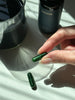 A woman's hand with long nails is holding a green capsule between her thumb and forefinger, and another capsule is resting on a table surface. There ia a glass of green liquid next to the capsule, along with a green bottle of juna detox capsules. 