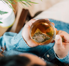 A mug of tea is shown from above. There is a clear mug with a teabag in it, and the tea is golden yellow. A woman's left palm holds the mug, and her right hand is on the handle. She is wearing a denim long sleeve shirt. Leaves of a plant are in the frame on the left.