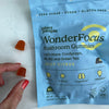 bag of wonderfocus mushroom gummies with lion's mane, cordyceps, b6, b12, and green tea. Bag is a light blue. Two orange gummies sit next to the bag and a woman's fingers are grabbing a gummy