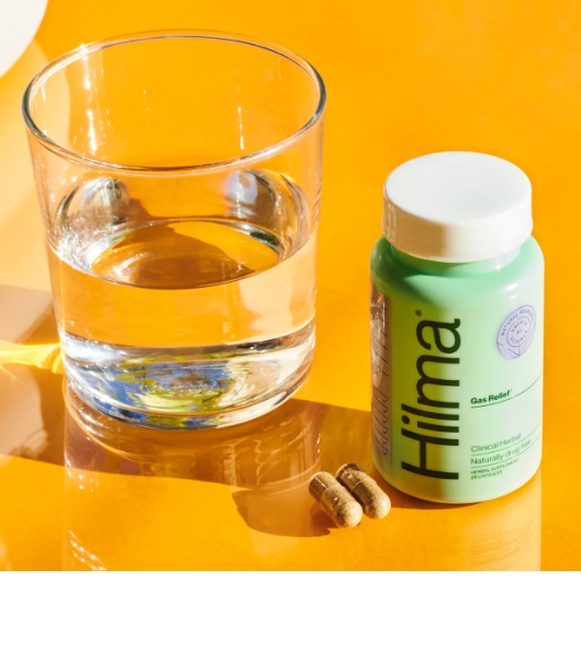 A clear half full glass of water against an orange background rests on a table next to 2 brown capsules, and a bottle of Hilma Gas Relief. The bottle is green with a white cap. 