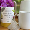 Peppermint chasteberry menopause tea next to a white mug, white teapot, and a vase of purple flowers. Two tea sachets sit next to the mug.