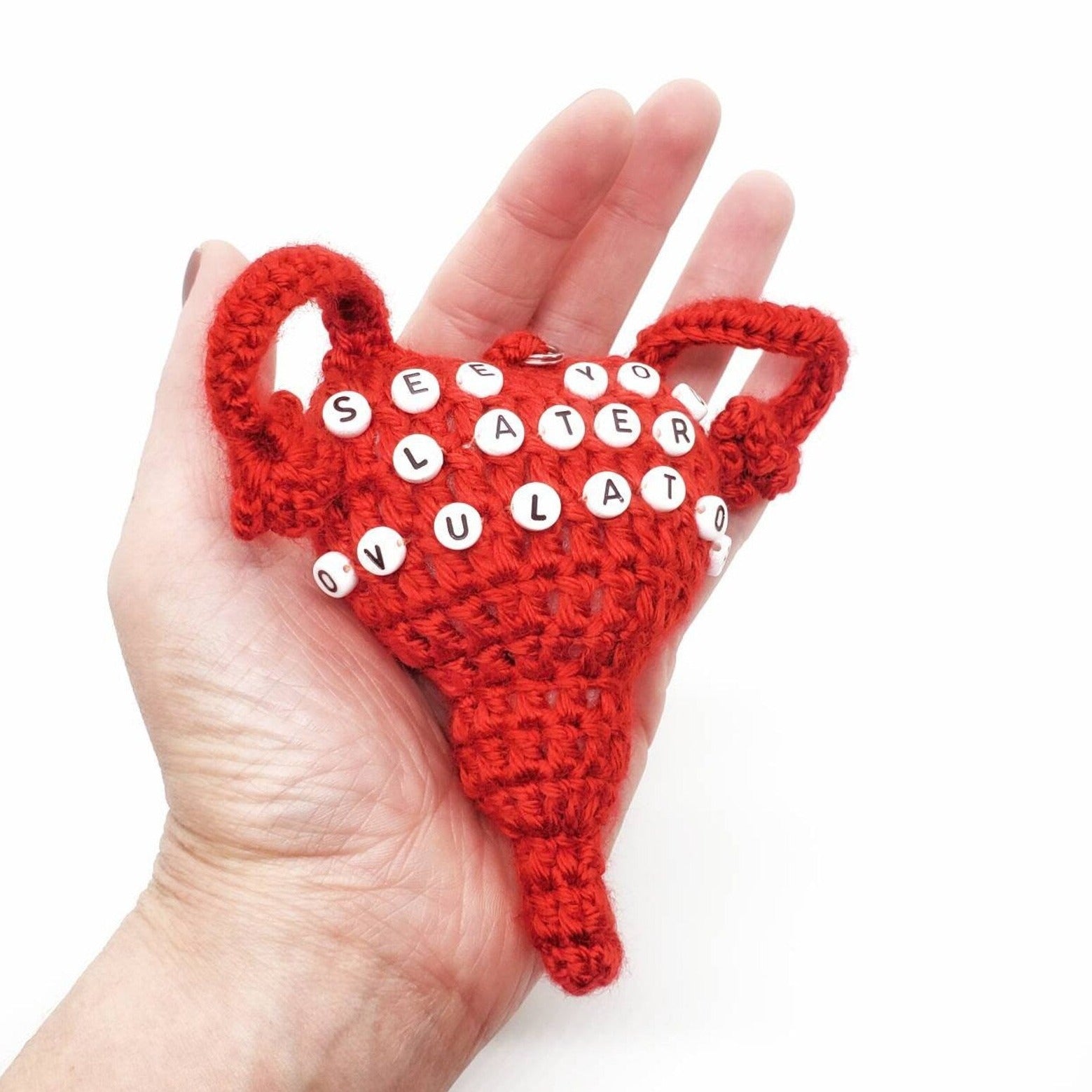 The palm of a hand holding a red acrylic yarn crocheted into the shape of a uterus with See You Later Ovulator spelled out in black and white beads.
